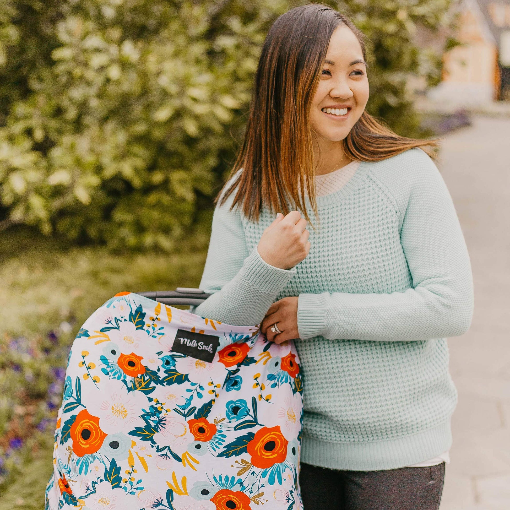 Woman holding an infant car seat with floral cover outdoors.