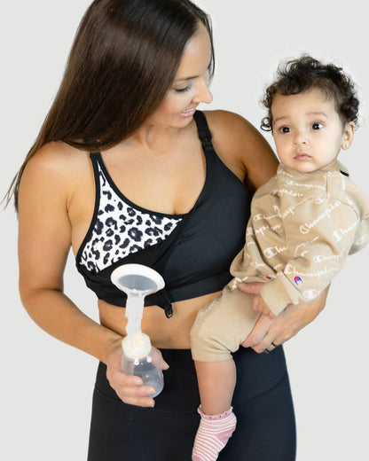 Woman holding a baby and a breast pump in a leopard print nursing and pumping sports bra against a white background