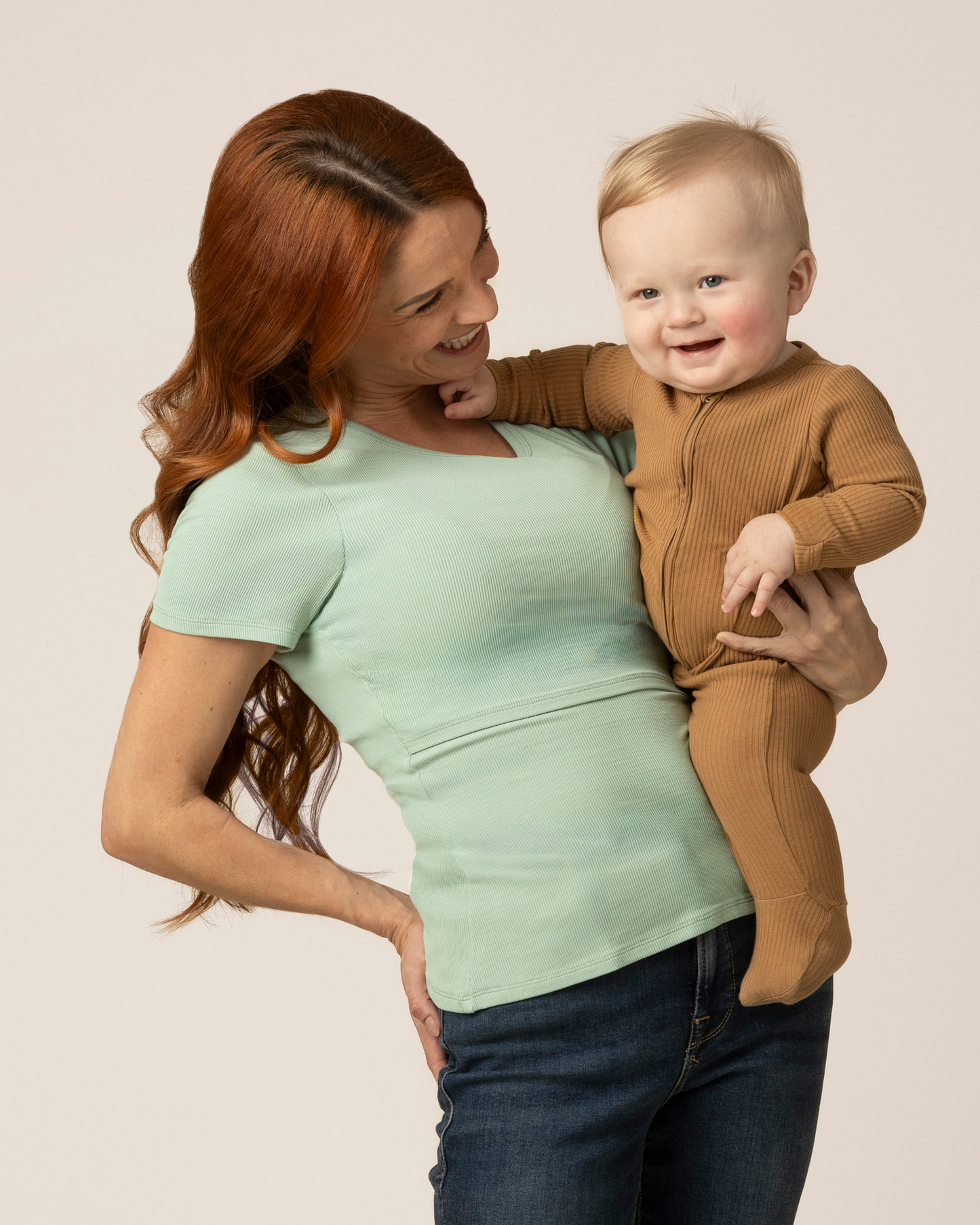 Woman wearing a green nursing top while holding a baby in a brown outfit against a plain background
