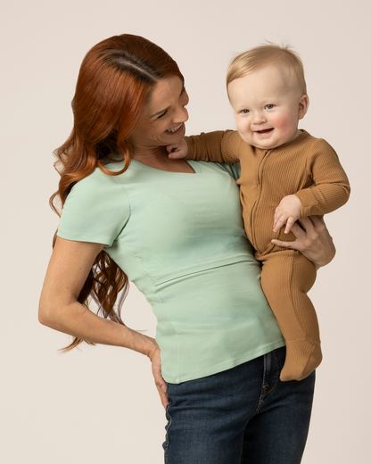 Woman wearing a green nursing top while holding a baby in a brown outfit against a plain background