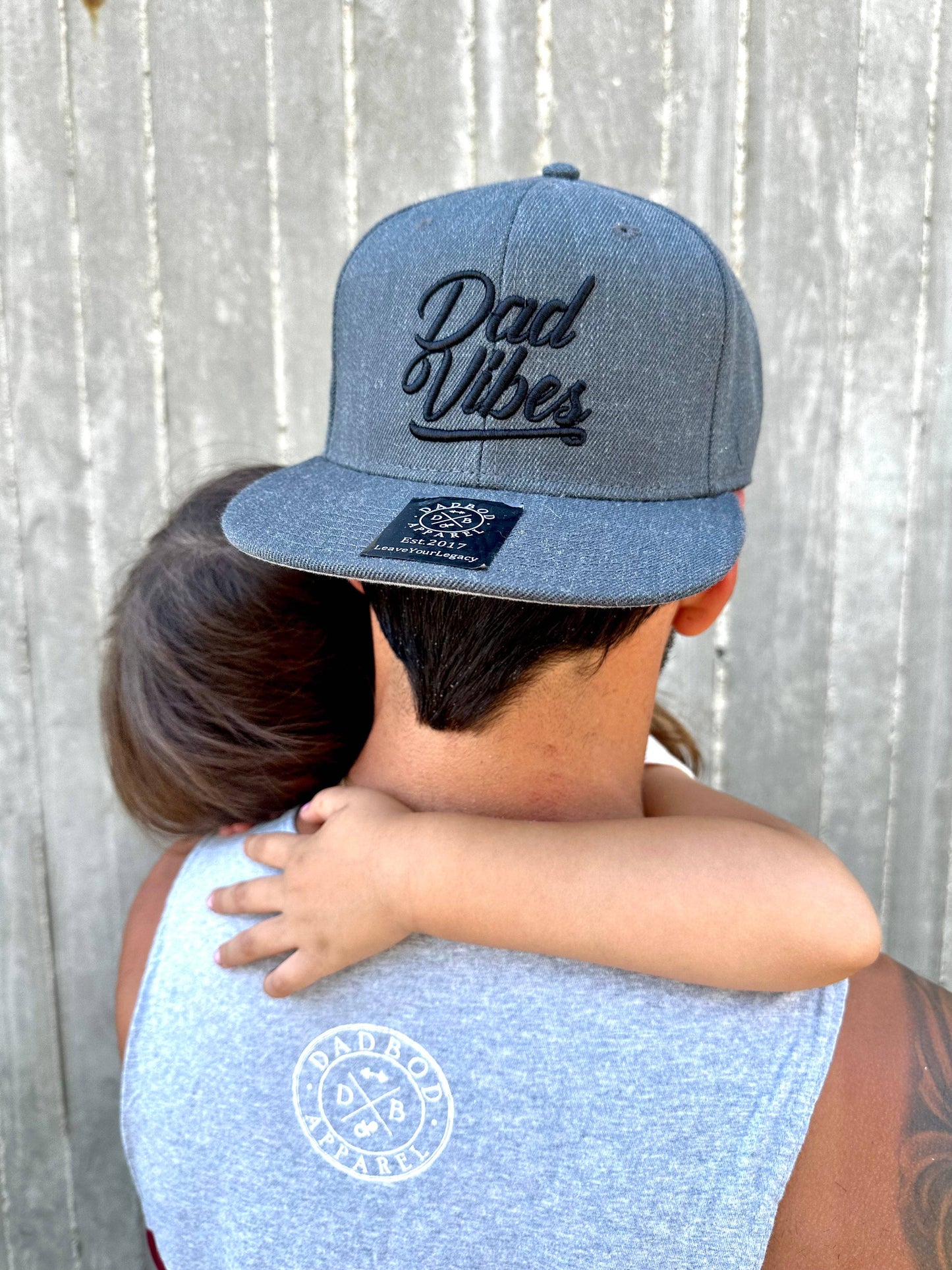 Person wearing a denim cap with 'Dad Vibes' text, embracing a child against a wooden background.