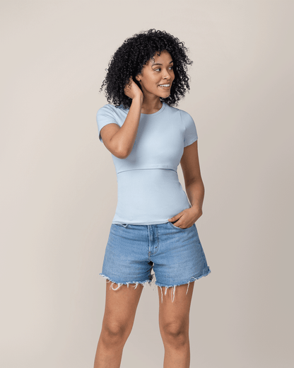 Woman wearing a light blue nursing t-shirt and denim shorts against a beige background