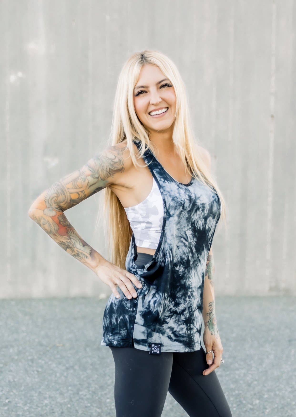 Woman wearing a tie-dye tank top and leggings with tattoos on her arms, standing against a plain background.