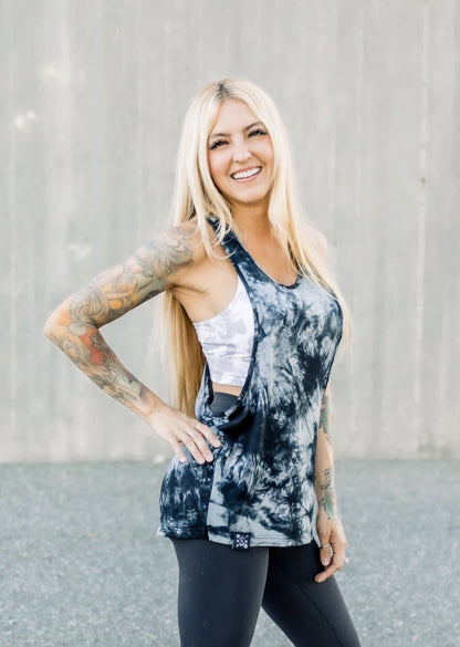 Woman wearing a tie-dye tank top and leggings with tattoos on her arms, standing against a plain background.