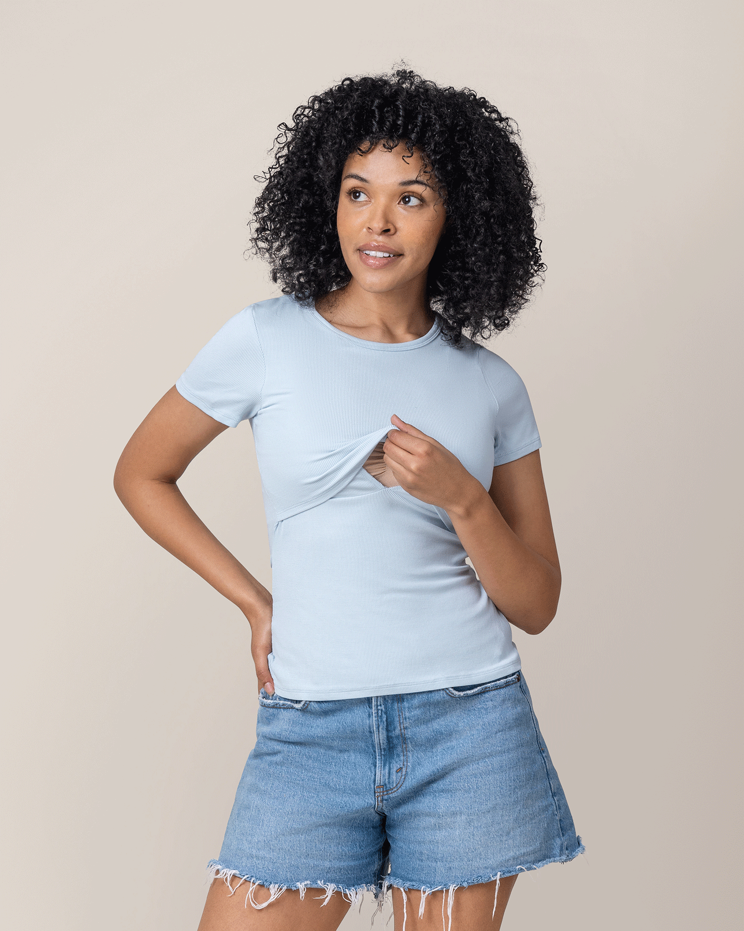 Woman wearing a light blue t-shirt with breastfeeding access and denim shorts against a beige background