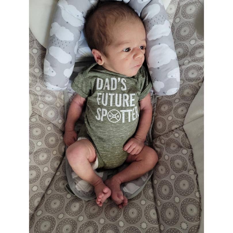 Baby wearing a green onesie with text that says "Dad's future spotter," sitting on a patterned surface.
