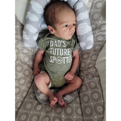 Baby wearing a green onesie with text that says "Dad's future spotter," sitting on a patterned surface.