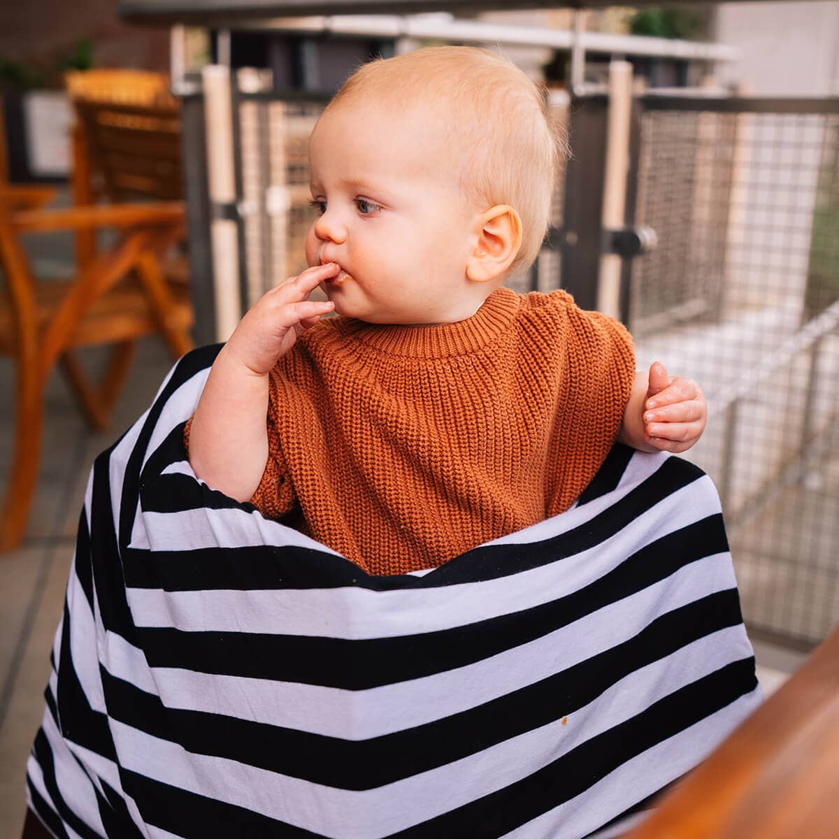 Baby sitting outdoors in a high chair with a black and white striped cover on the chair with an orange sweater.