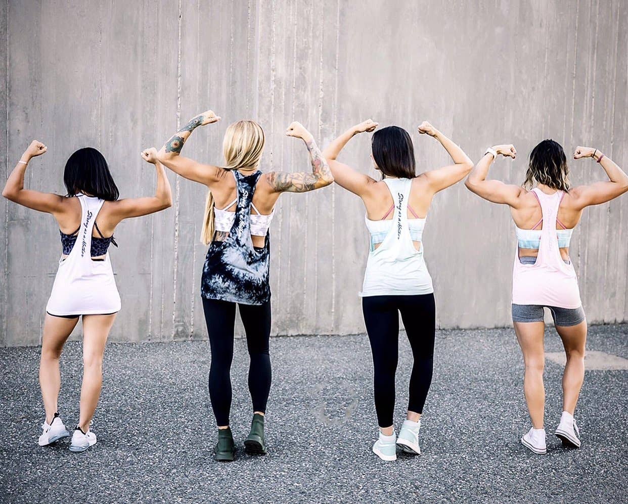 Four women in athletic wear flexing their backs and arms against a concrete wall.