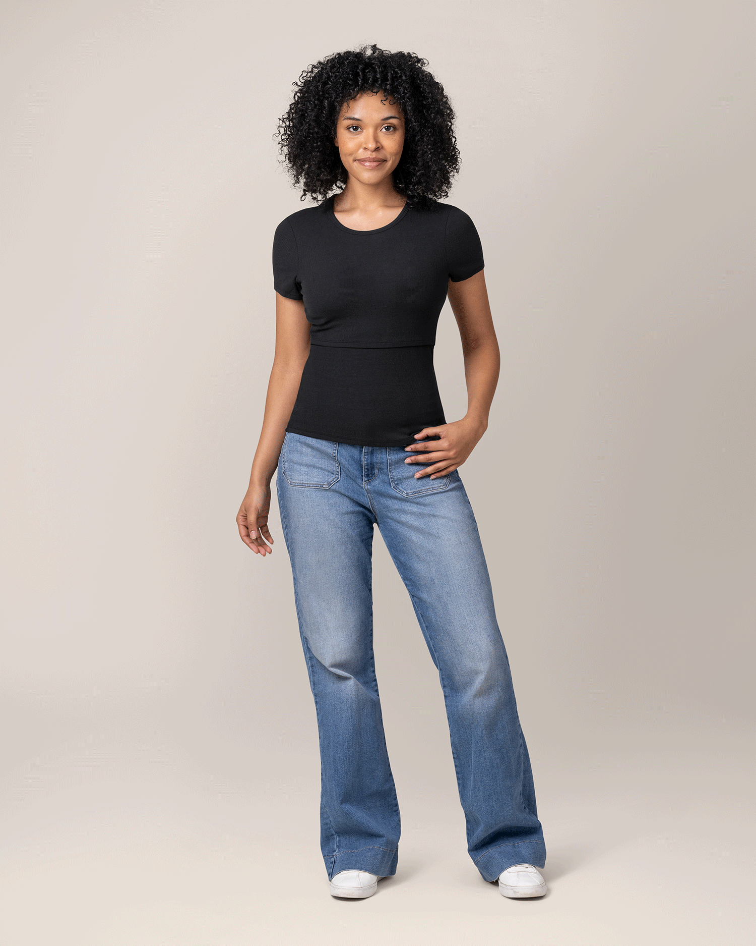 Woman wearing a black, ribbed t-shirt with nursing access and blue jeans on a plain background