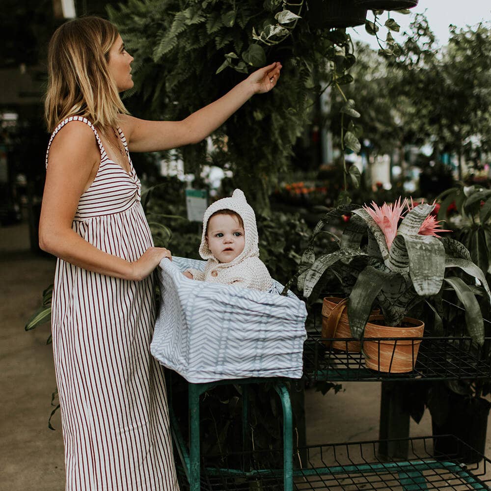 Woman in a striped dress holding a baby in a basket among plants with a blue patterned car seat cover.