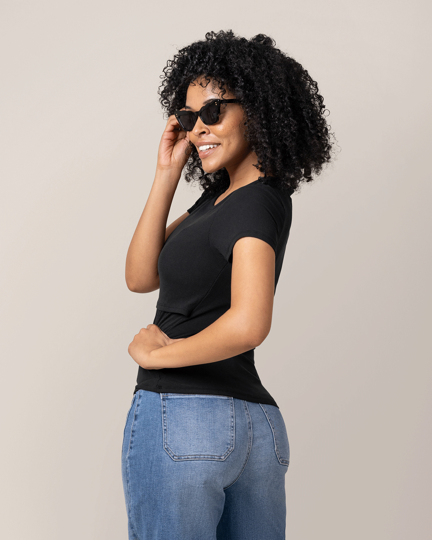 Woman wearing a black top and blue jeans with sunglasses on a plain background