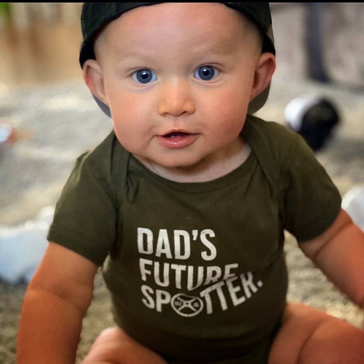 Baby wearing a green onesie with 'Dad's Future Spotter' text, sitting on a carpeted floor.
