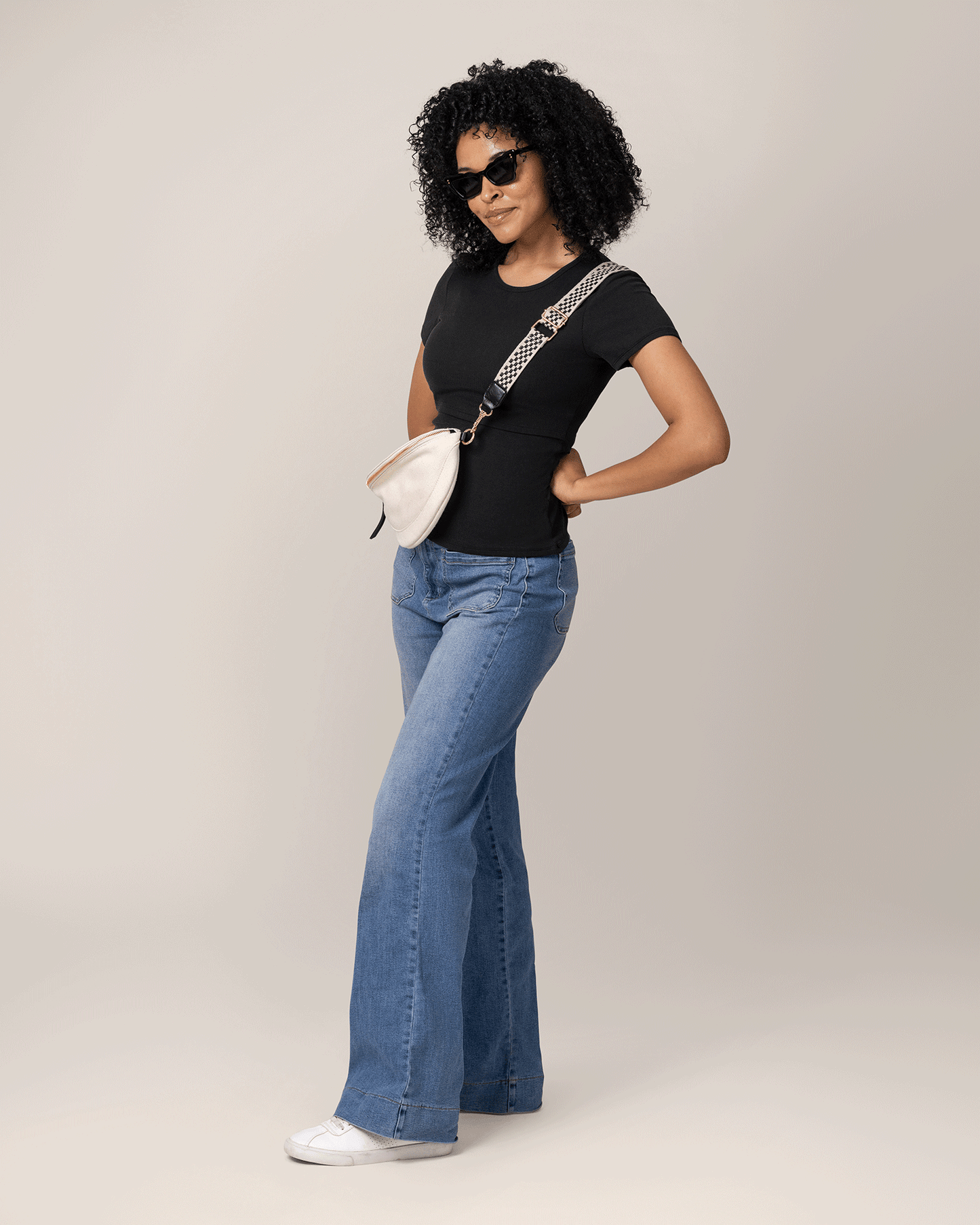 Woman wearing a black nursing t-shirt, blue jeans, and sunglasses with a white bag on a plain background